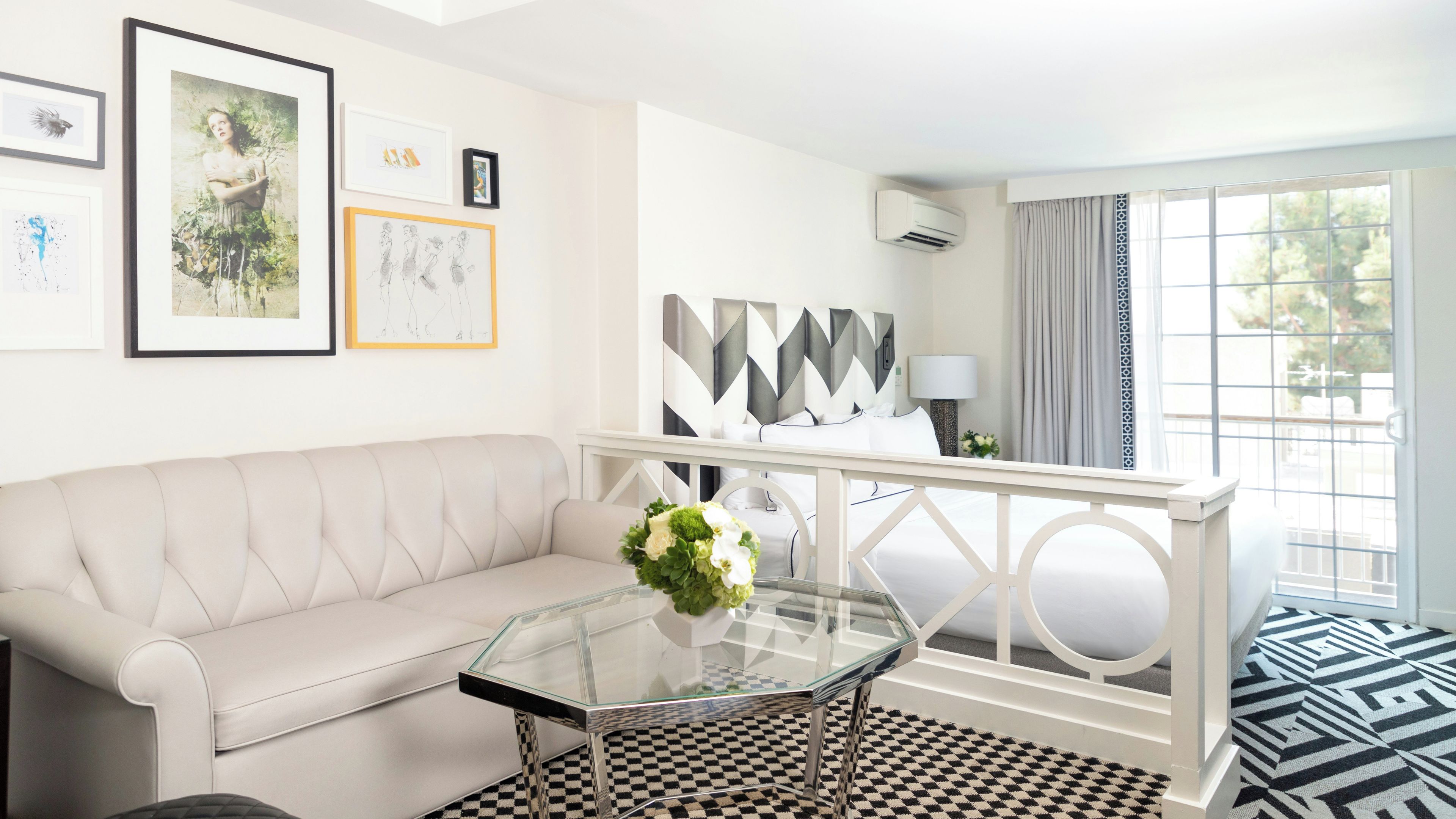 A modern hotel room with a white sofa, a glass coffee table with a flower arrangement, a geometric-patterned rug, and a view of the outdoors through a large window.