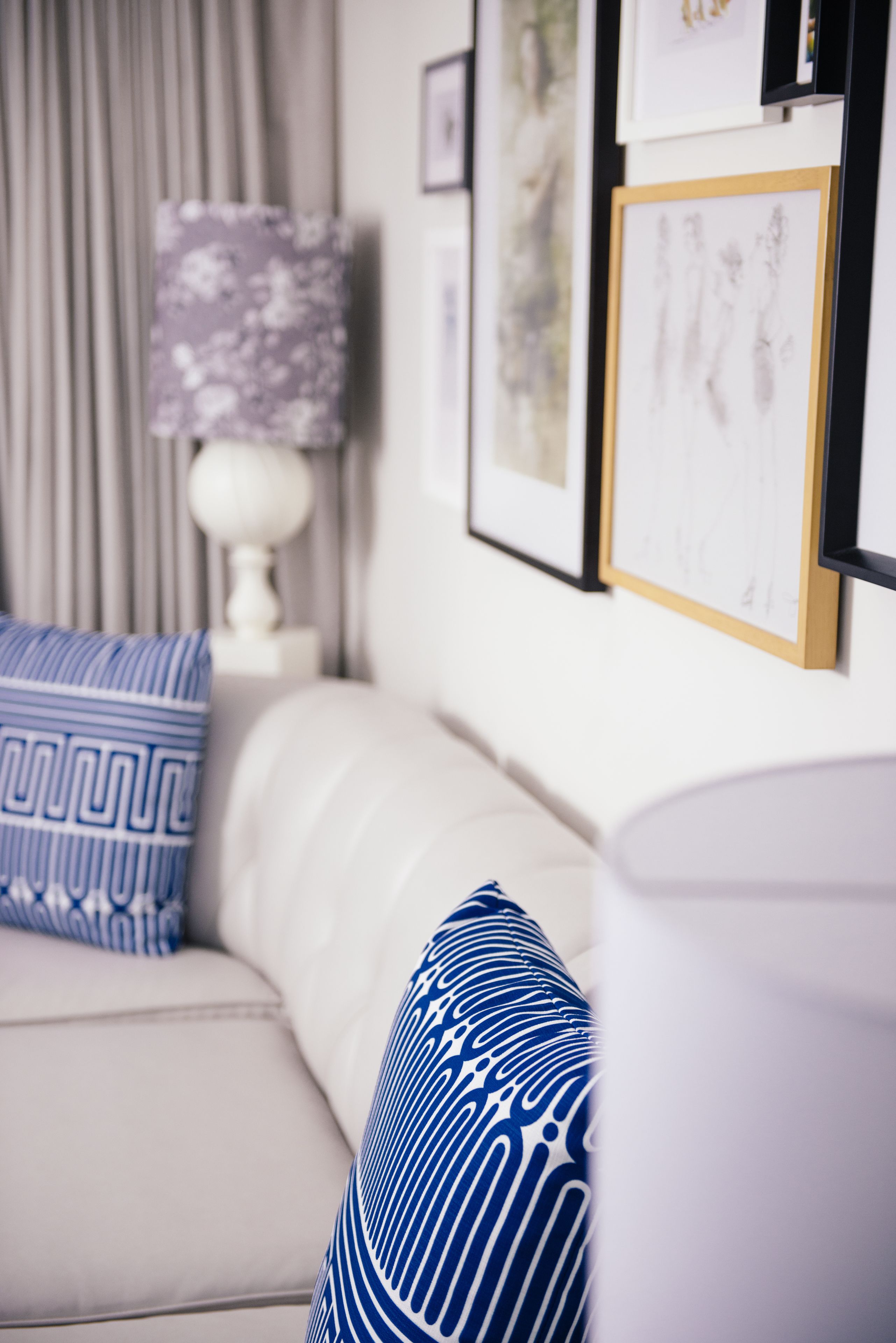 A living room with a white sofa, blue patterned pillows, a white lamp, and framed artwork on the wall.