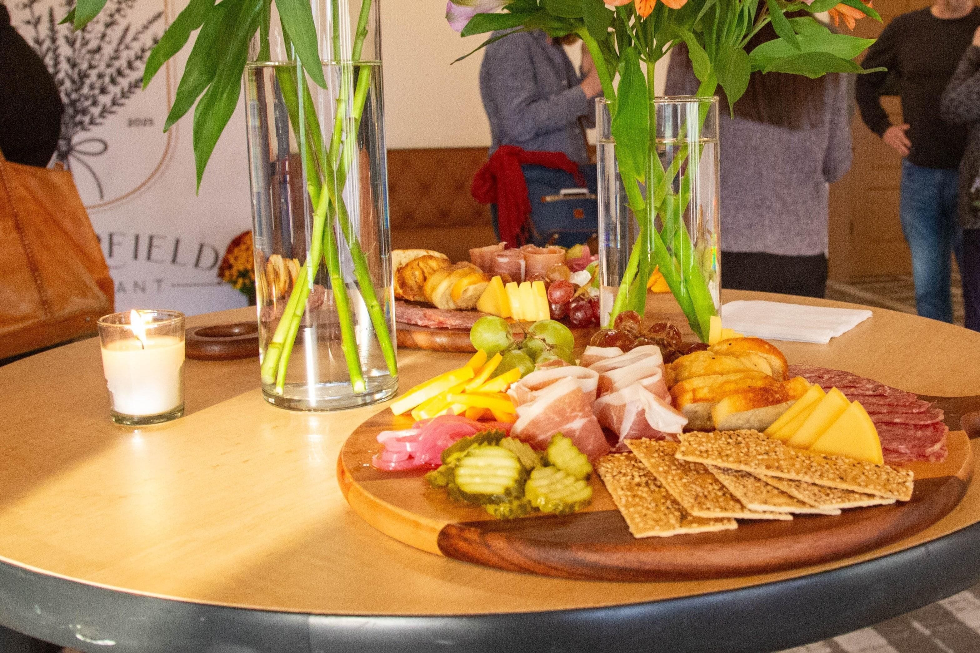 A wooden table with a charcuterie board, candles, and vases with green plants.