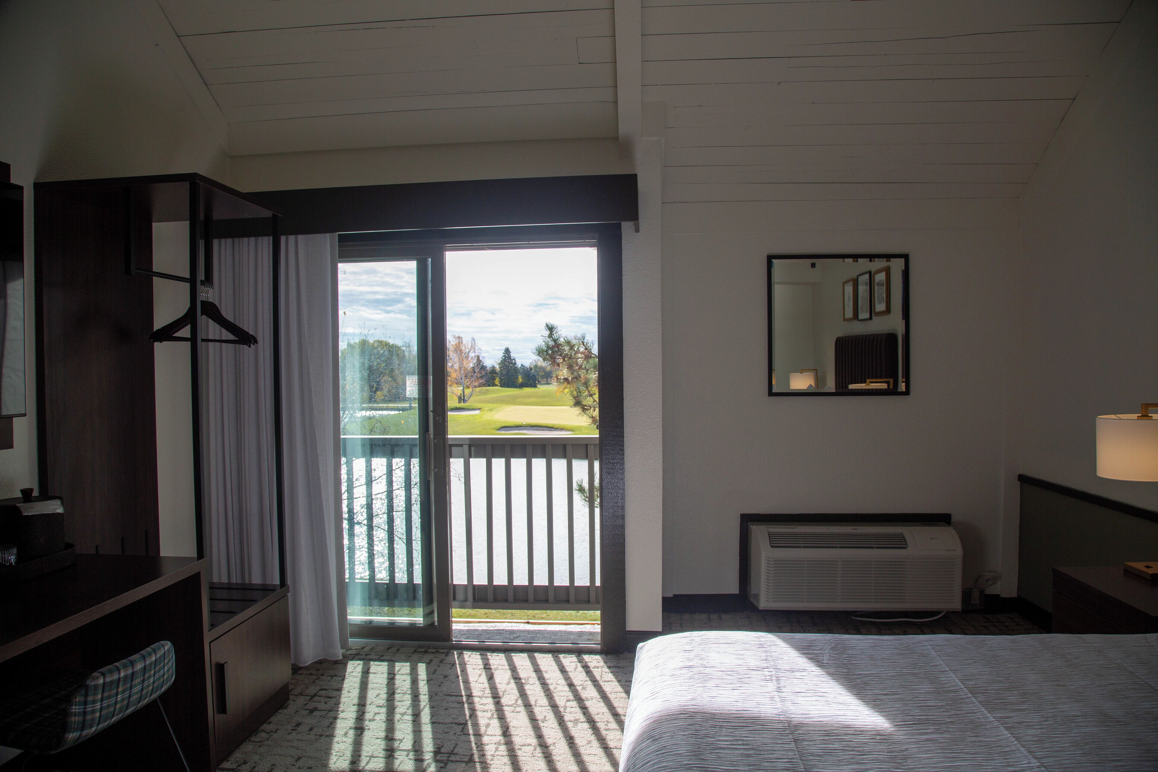 A hotel room with a bed, a desk, a chair, a hanging coat rack, a window with a view of a golf course, and a wall-mounted mirror.