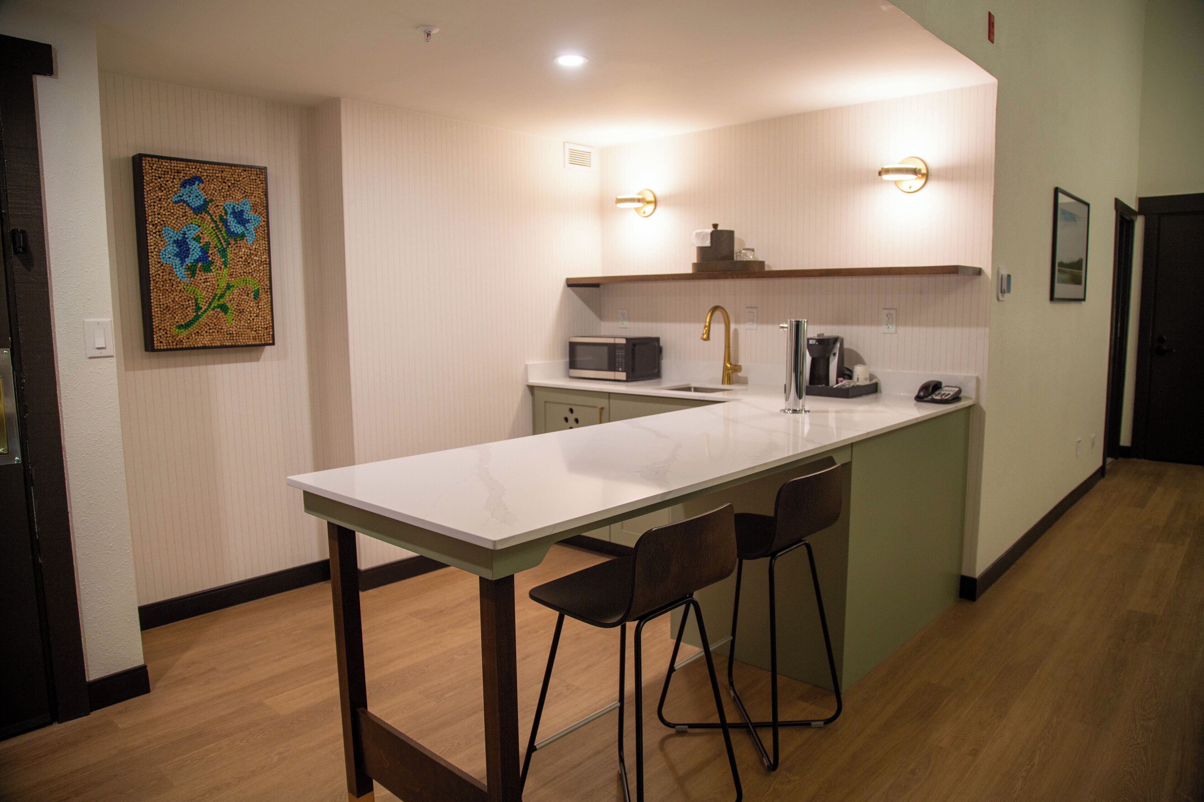 A modern kitchen with a white countertop, two bar stools, a microwave, a coffee maker, and a sink.