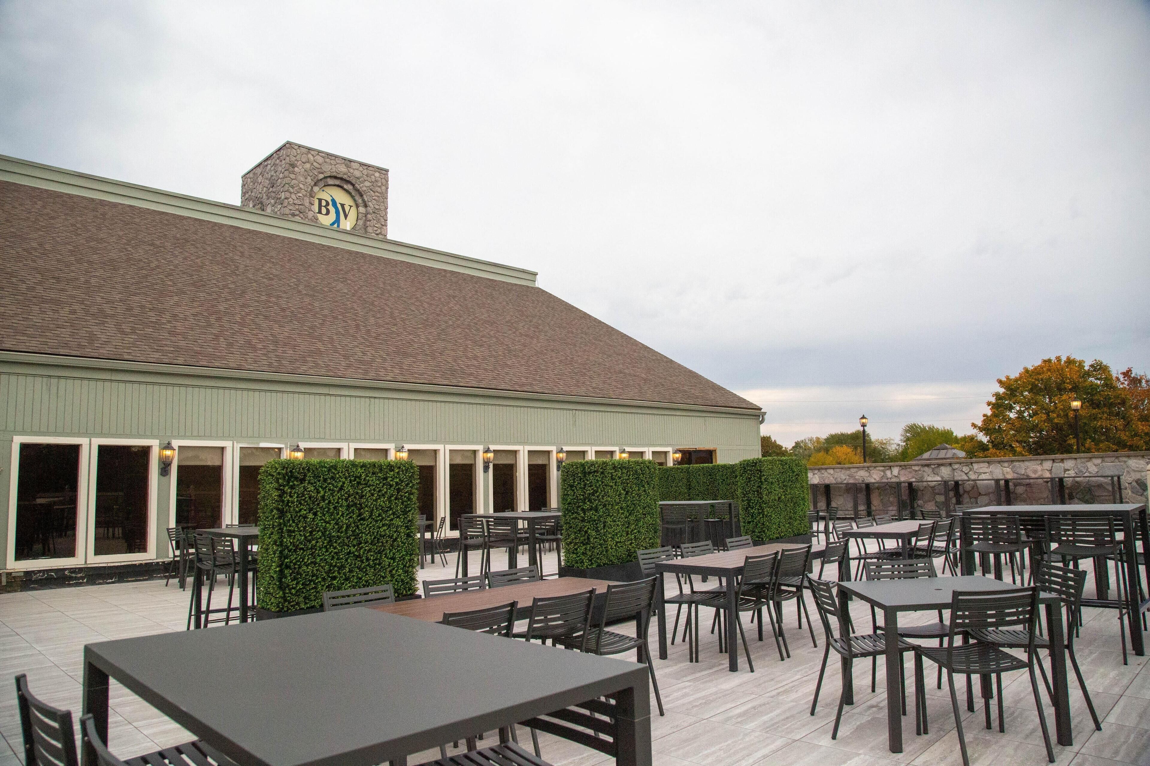 An outdoor patio with tables and chairs, a stone wall, and a building with a 'BV' logo.