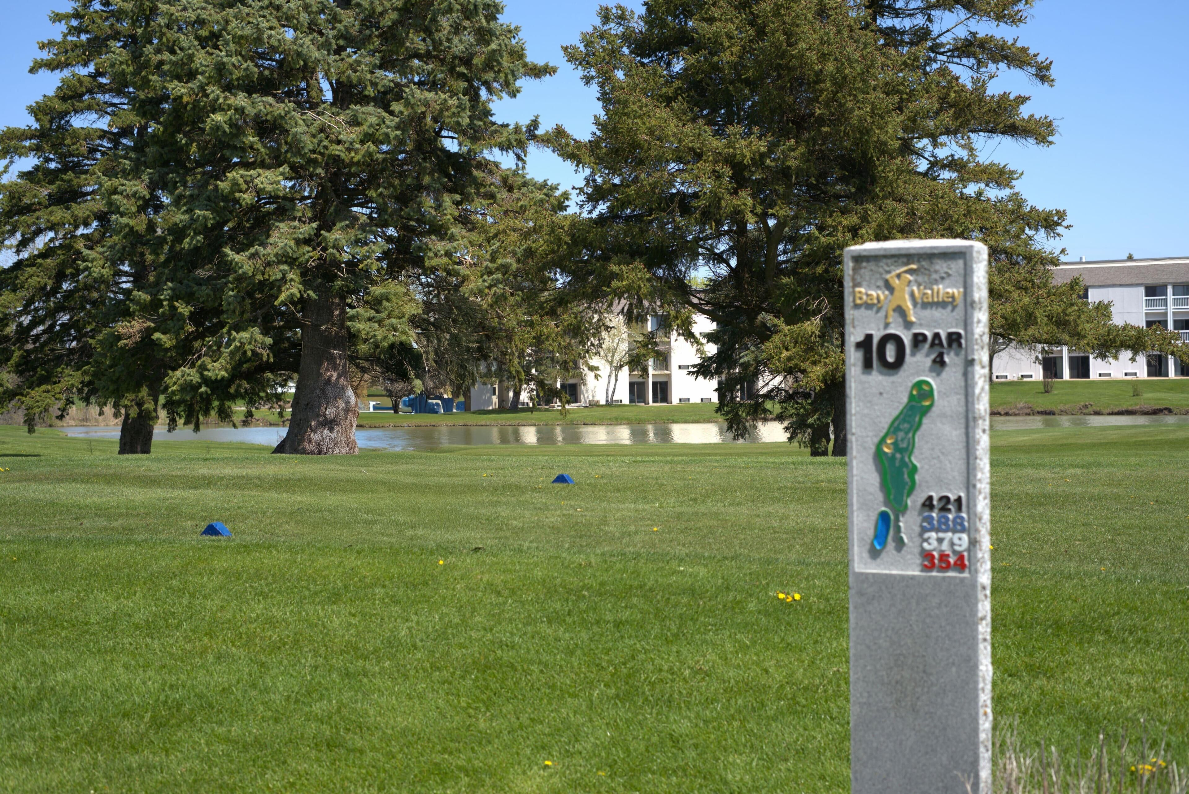 A golf course with a sign indicating a 10 par hole, surrounded by trees and a building in the background.