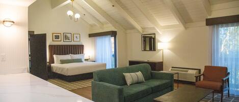 A hotel room with a bed, a green sofa, a small table, and a chair. There are ceiling lights and a window with curtains.
