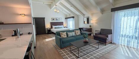 Una habitación de hotel moderna con cocina, sala de estar con sofá y mesa de centro, comedor independiente y un ventanal con cortinas.