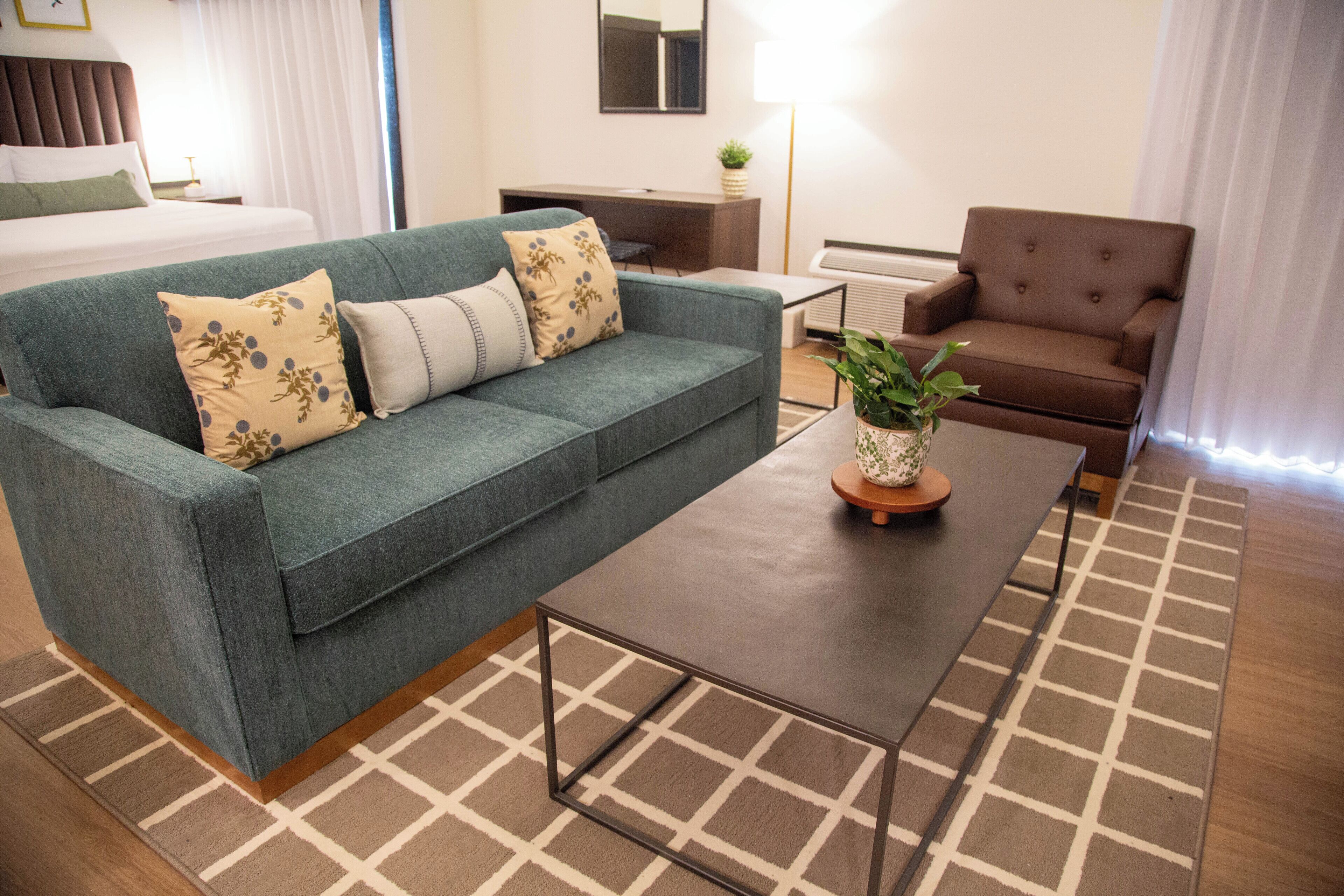 Una habitación de hotel moderna con un sofá verde, una mesa de centro de madera, un sillón marrón y una cama con ropa de cama blanca.