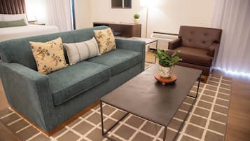 Una habitación de hotel moderna con un sofá verde, una mesa de centro de madera, un sillón marrón y una cama con ropa de cama blanca.