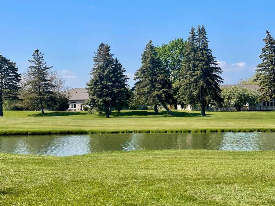 Un campo de golf bien cuidado con un estanque, rodeado de altos árboles perennes y un edificio al fondo.