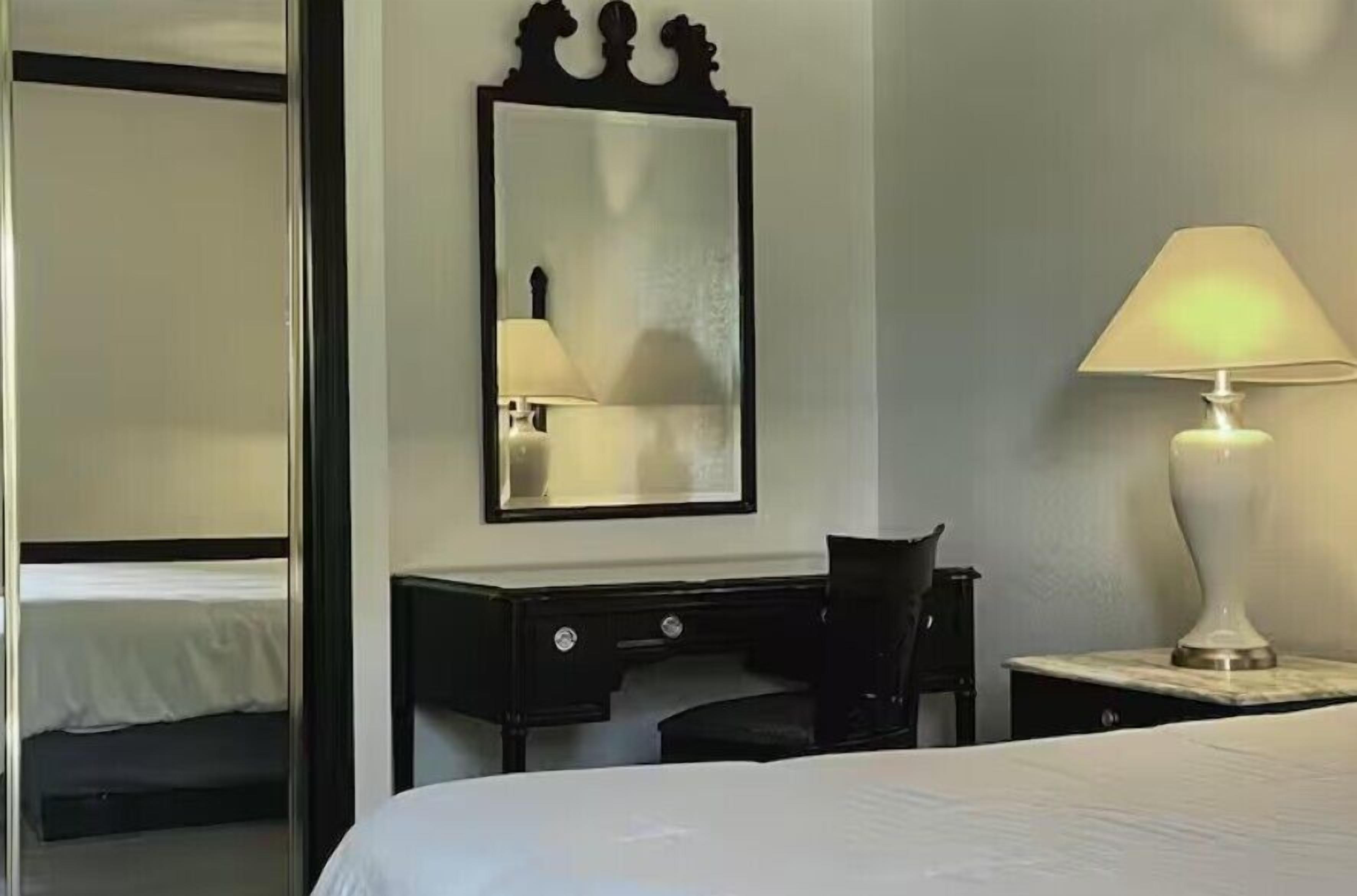 A hotel room with a bed, a desk, a chair, a lamp, and a mirror.