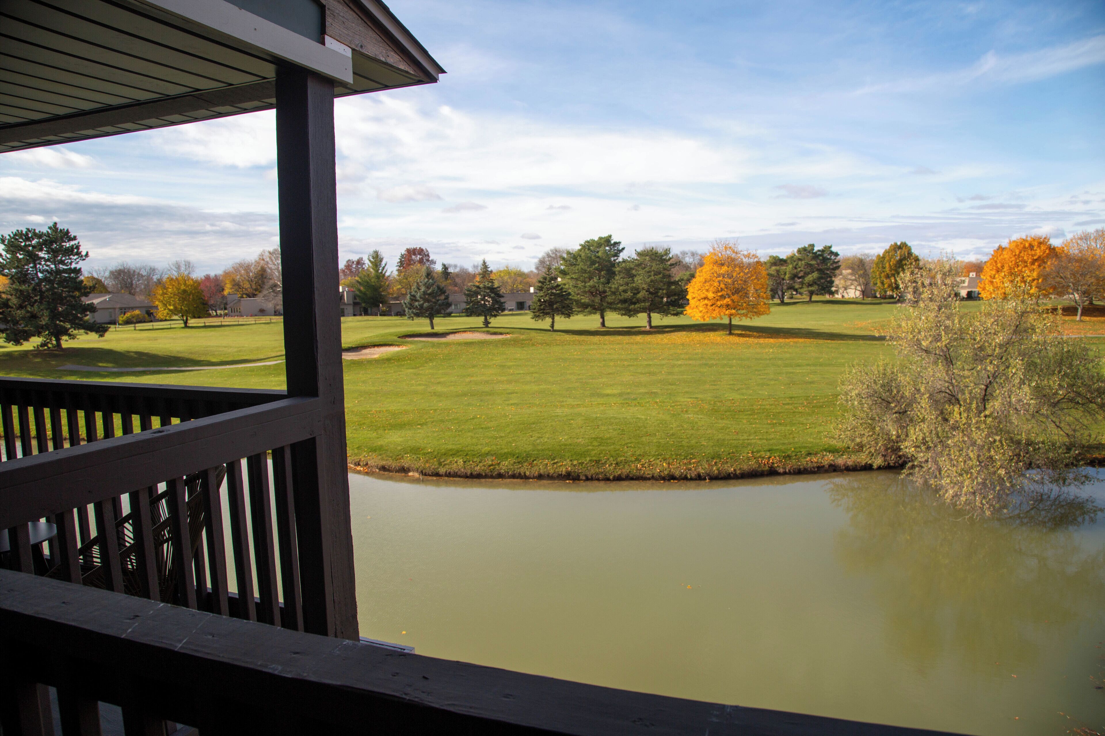 A view from a covered deck overlooking a grassy area with a pond and trees in autumn colors.