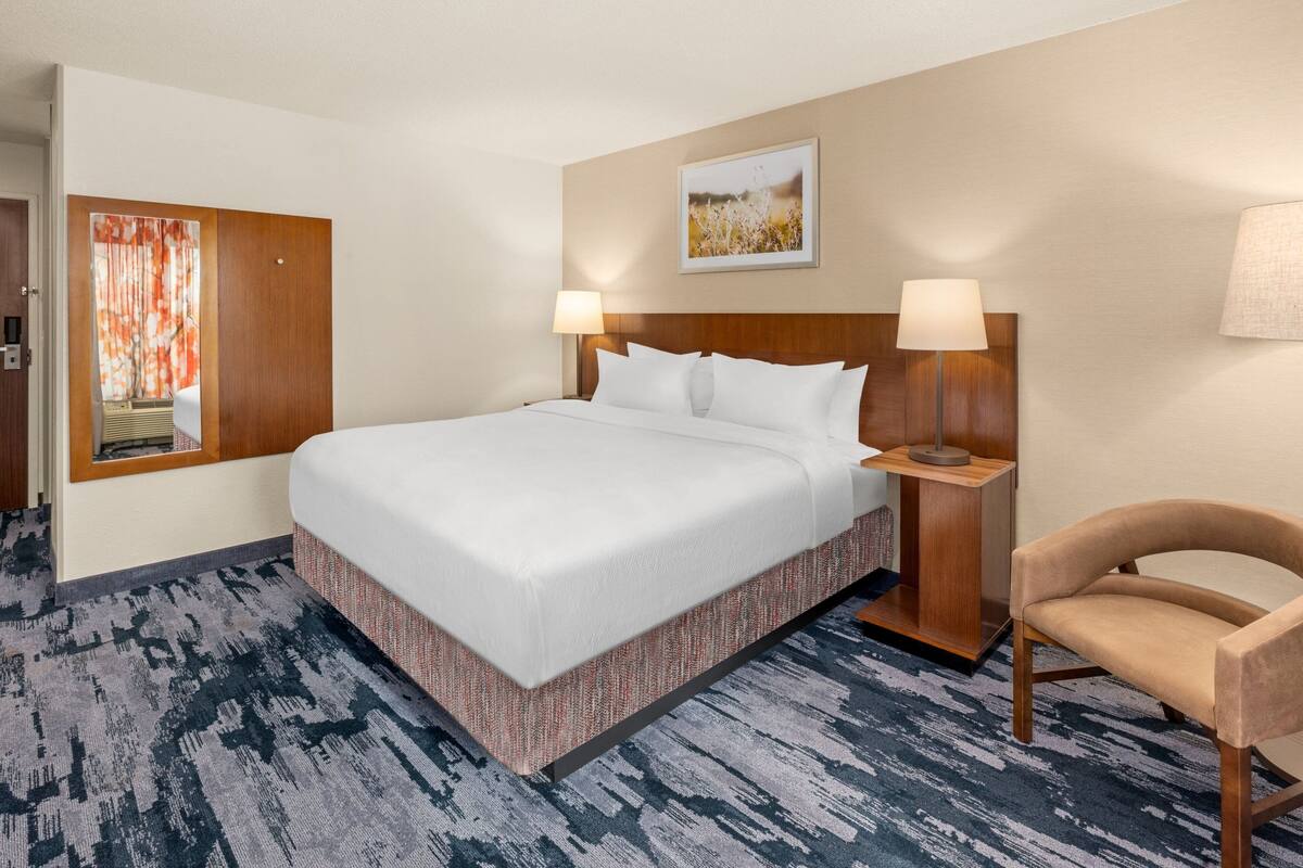 A hotel room with a large bed, two bedside lamps, a wooden headboard, a chair, and a painting on the wall.