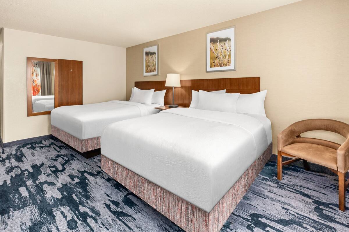 A hotel room with two beds, a wooden headboard, a mirror, a lamp, and a chair.
