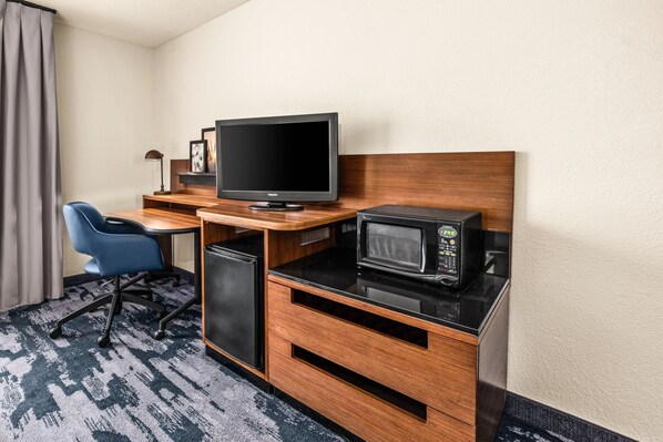 A compact hotel room with a wooden desk, a television, a microwave, a small refrigerator, and a blue chair.