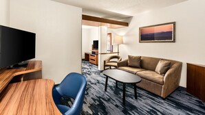 A hotel room with a flat-screen TV, a sofa, a blue chair, a small table, and a painting on the wall.