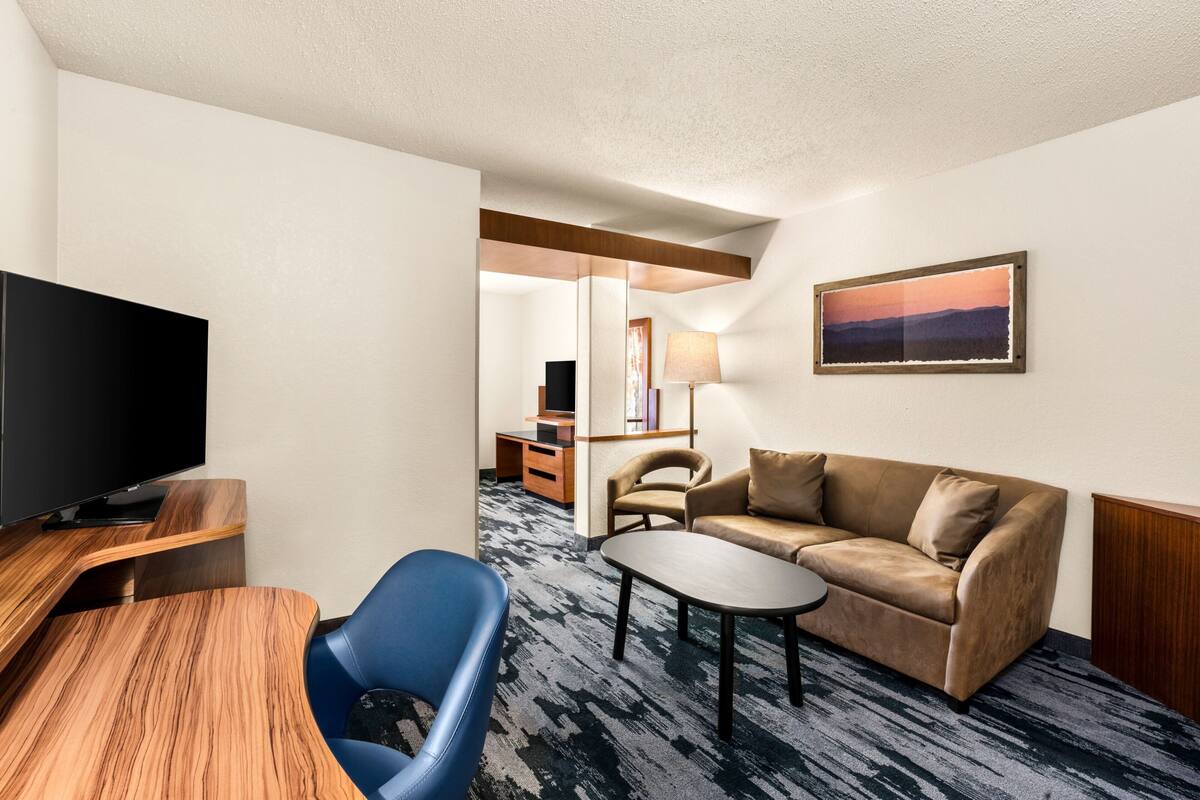 A hotel room with a flat-screen TV, a sofa, a blue chair, a small table, and a painting on the wall.