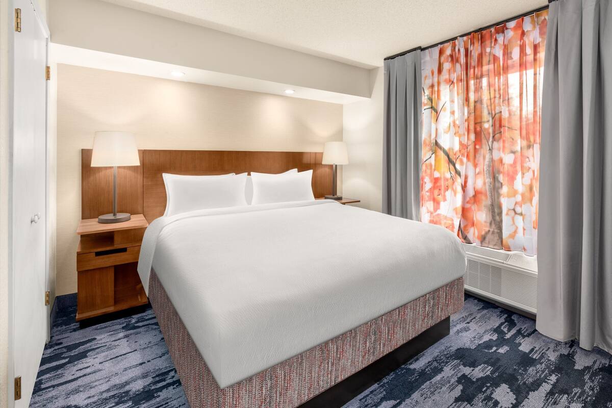 A hotel room with a large bed, two bedside tables with lamps, a window with curtains, and a carpeted floor.