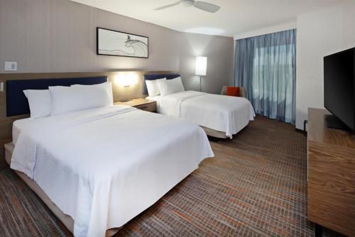 Ein Hotelzimmer mit zwei Betten, einem Deckenventilator, einem Flachbildfernseher und einem Fenster mit Vorhängen.