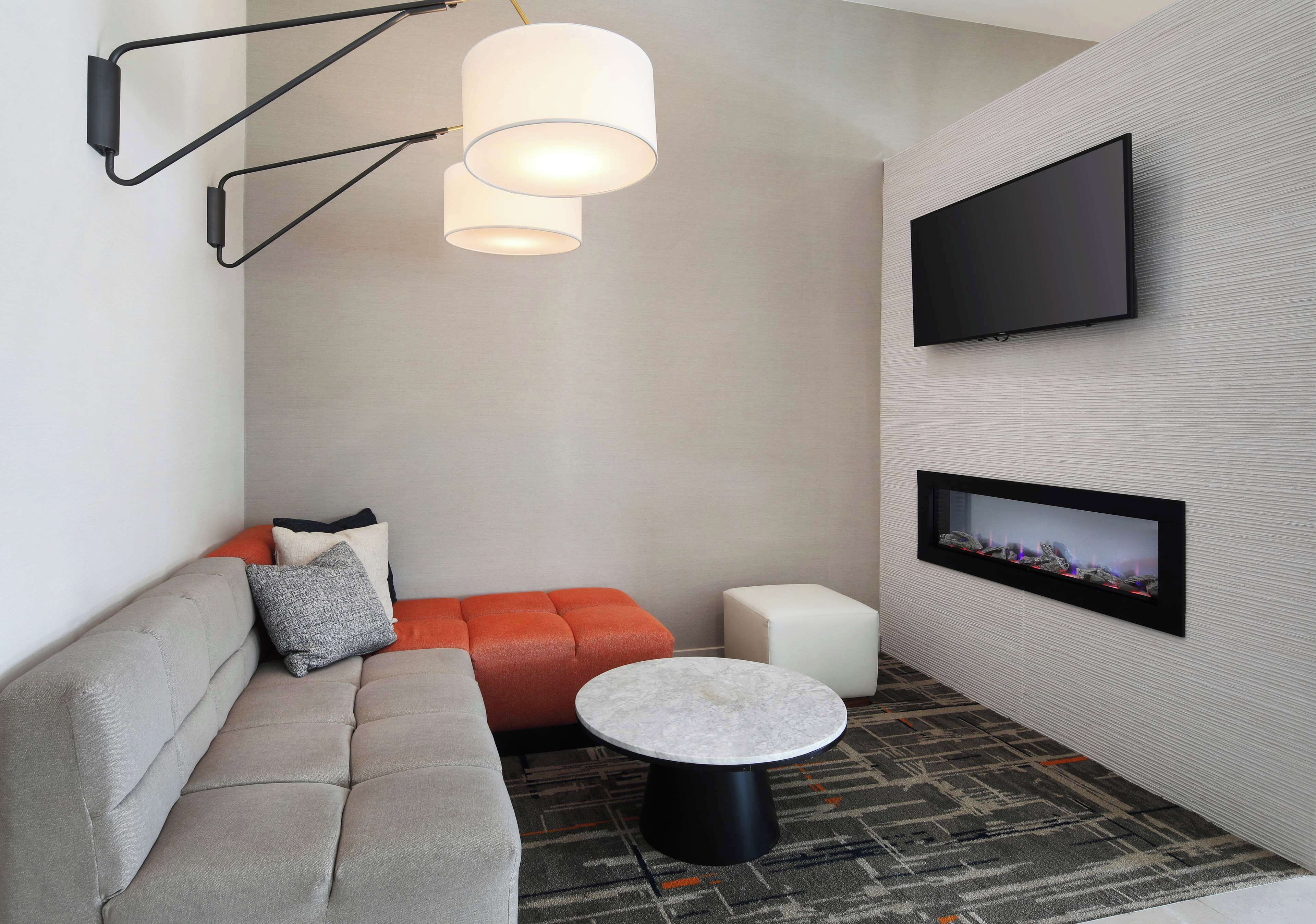 A modern living room with a grey sofa, orange accent seating, a round coffee table, a flat-screen TV mounted on the wall, and a patterned carpet.