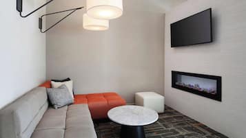A modern living room with a grey sofa, orange accent seating, a round coffee table, a flat-screen TV mounted on the wall, and a patterned carpet.
