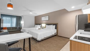 A hotel room with two beds, a dining table, a kitchen area, and a large window with curtains.