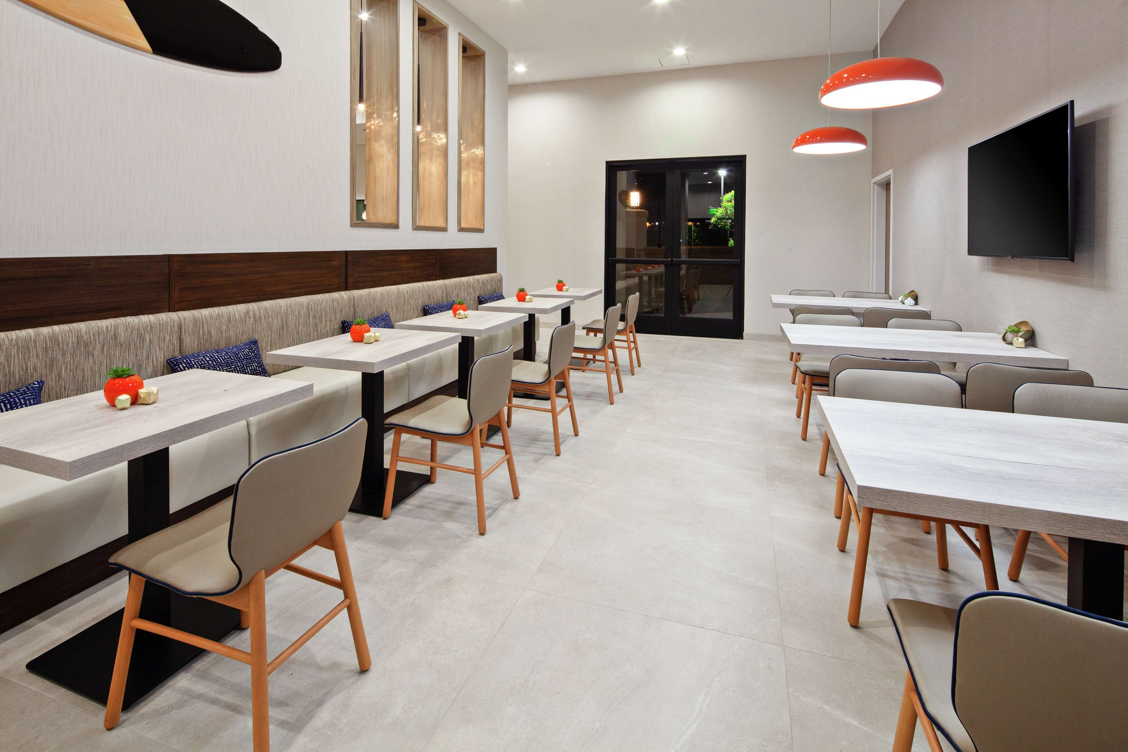 A modern dining area with multiple tables and chairs, a large TV mounted on the wall, and a bench along the wall.