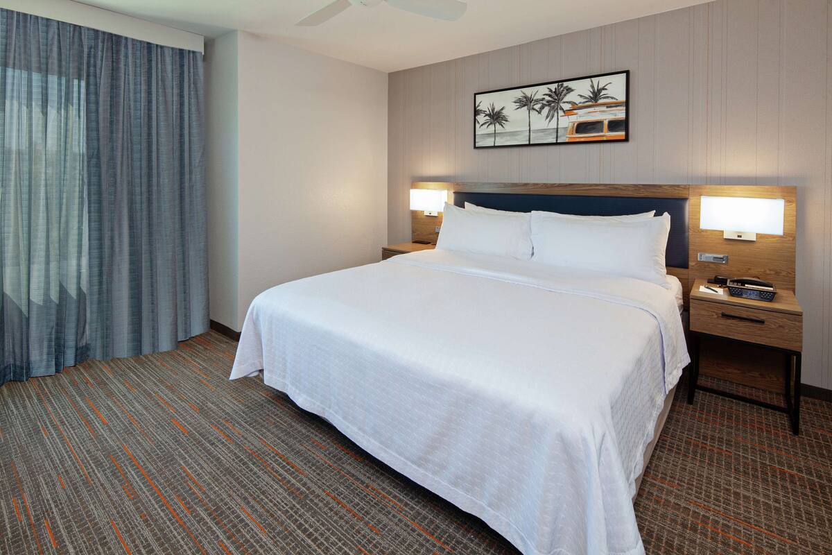 Une chambre d’hôtel avec un grand lit, deux tables de chevet, une petite table d’appoint, un ventilateur de plafond et un tableau représentant des palmiers au mur.
