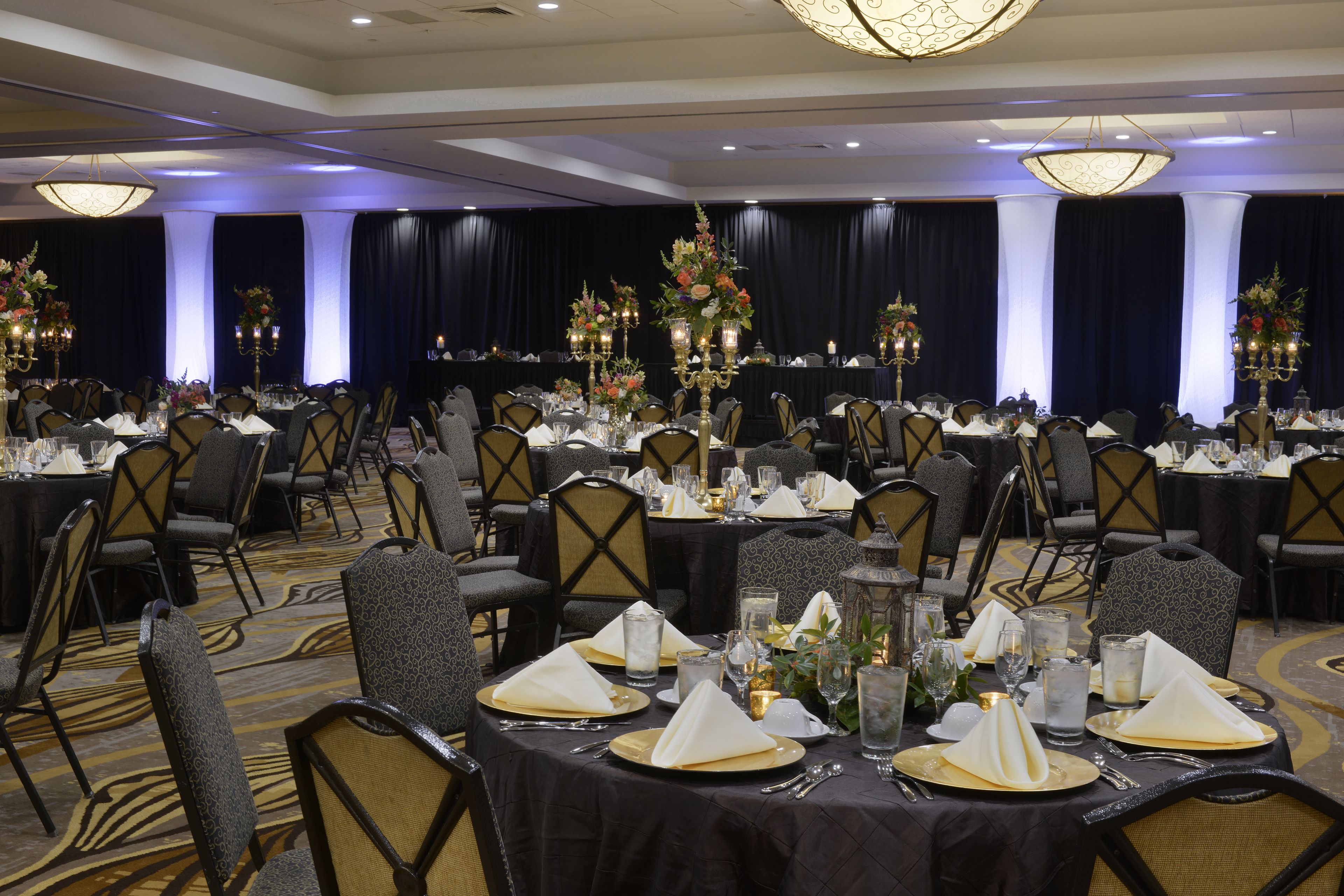 A banquet hall with round tables set for a formal event, featuring gold and black tablecloths, white napkins, and floral centerpieces.