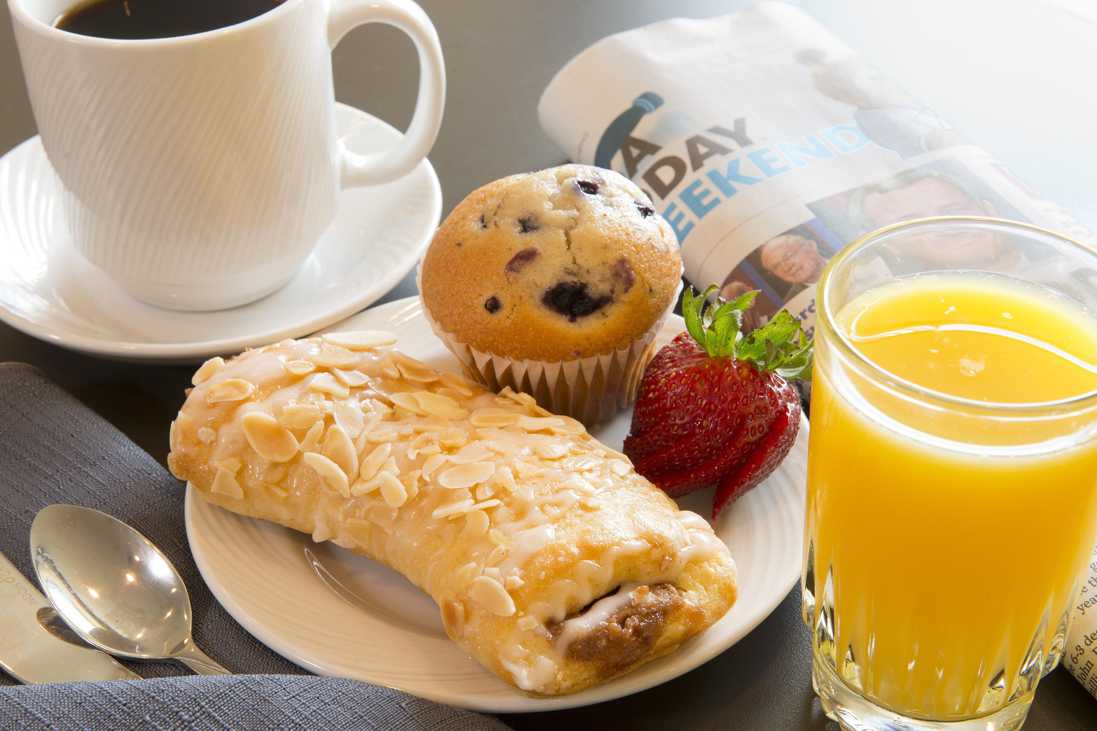 Mesa de café da manhã com muffin, pastel, copo de suco de laranja, xícara de café e jornal.