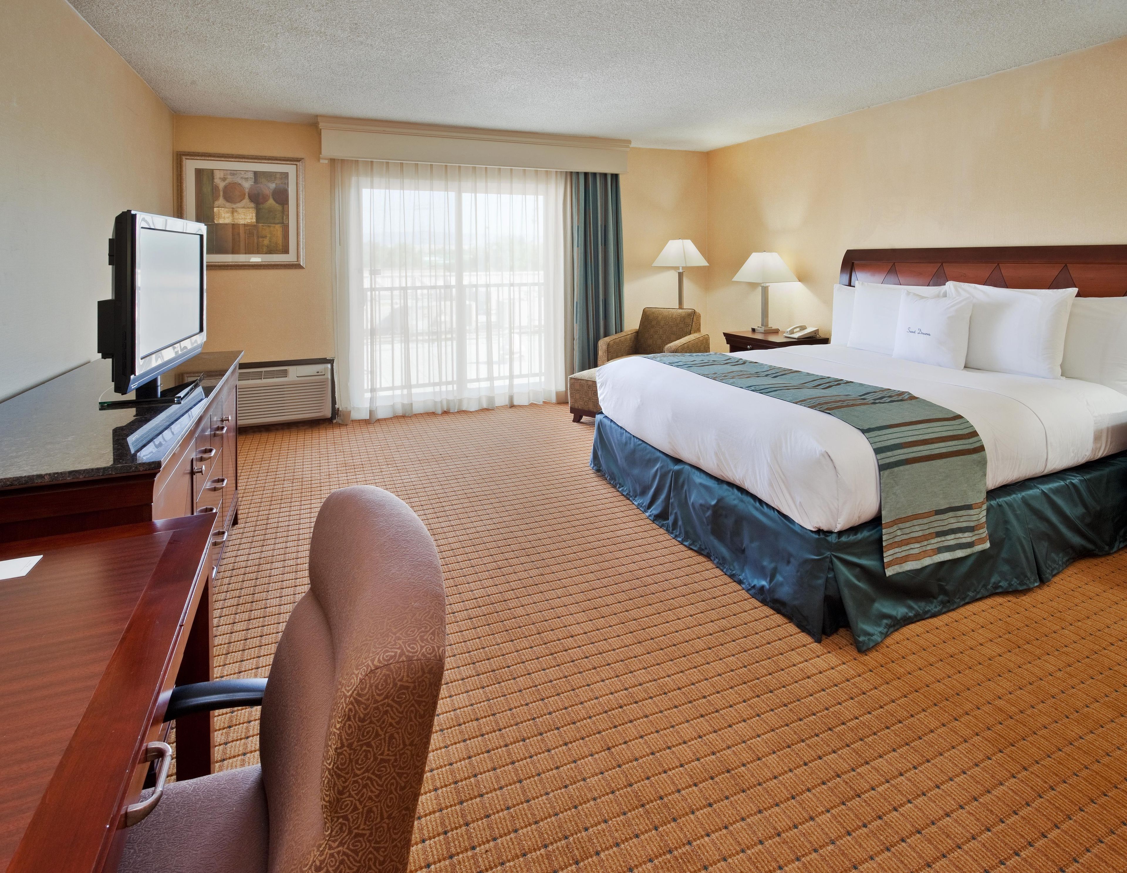 A hotel room with a bed, a desk with a television, a chair, a window with curtains, and a nightstand with lamps.