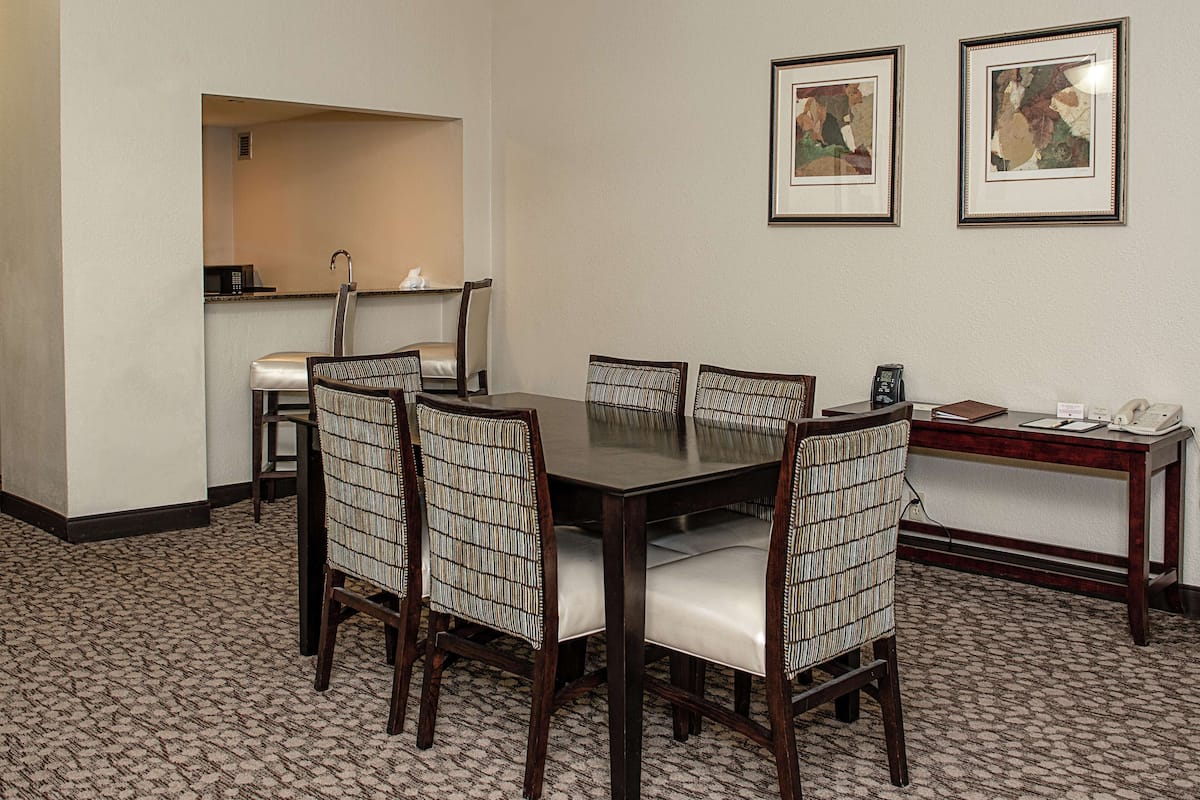 A dining area with a rectangular table, six chairs with cushioned seats, a small bar area with a microwave, and two framed artworks on the wall.