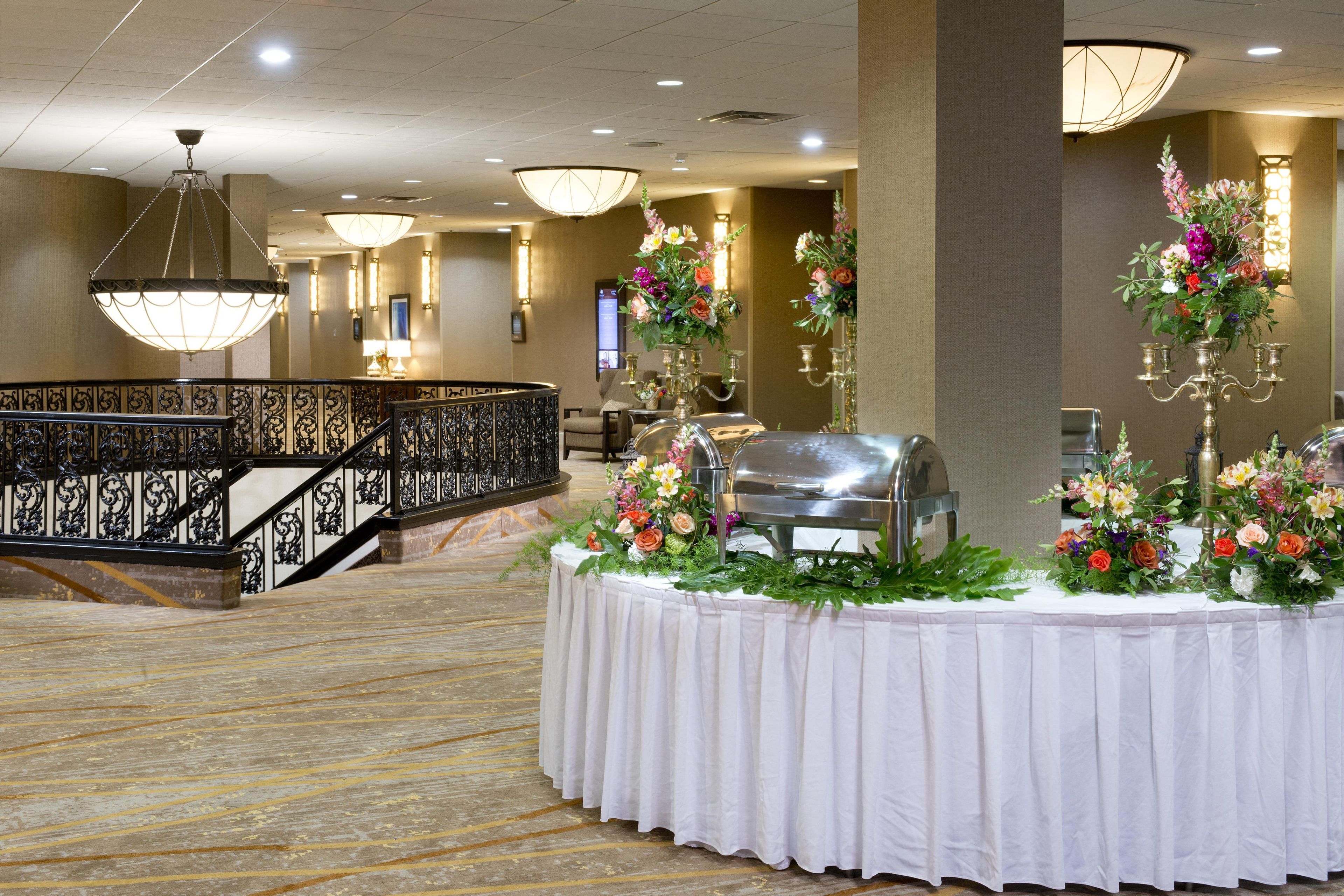 A hotel lobby with a reception desk, a buffet area with floral centerpieces, and a staircase with ornate railings.