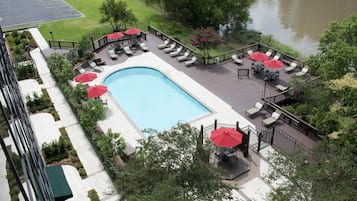 A rectangular swimming pool with lounge chairs and red umbrellas, surrounded by a deck with a view of a river.