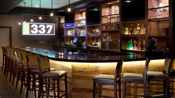 A bar area with a long counter, multiple bar stools, shelves with bottles, and a TV mounted above the bar.