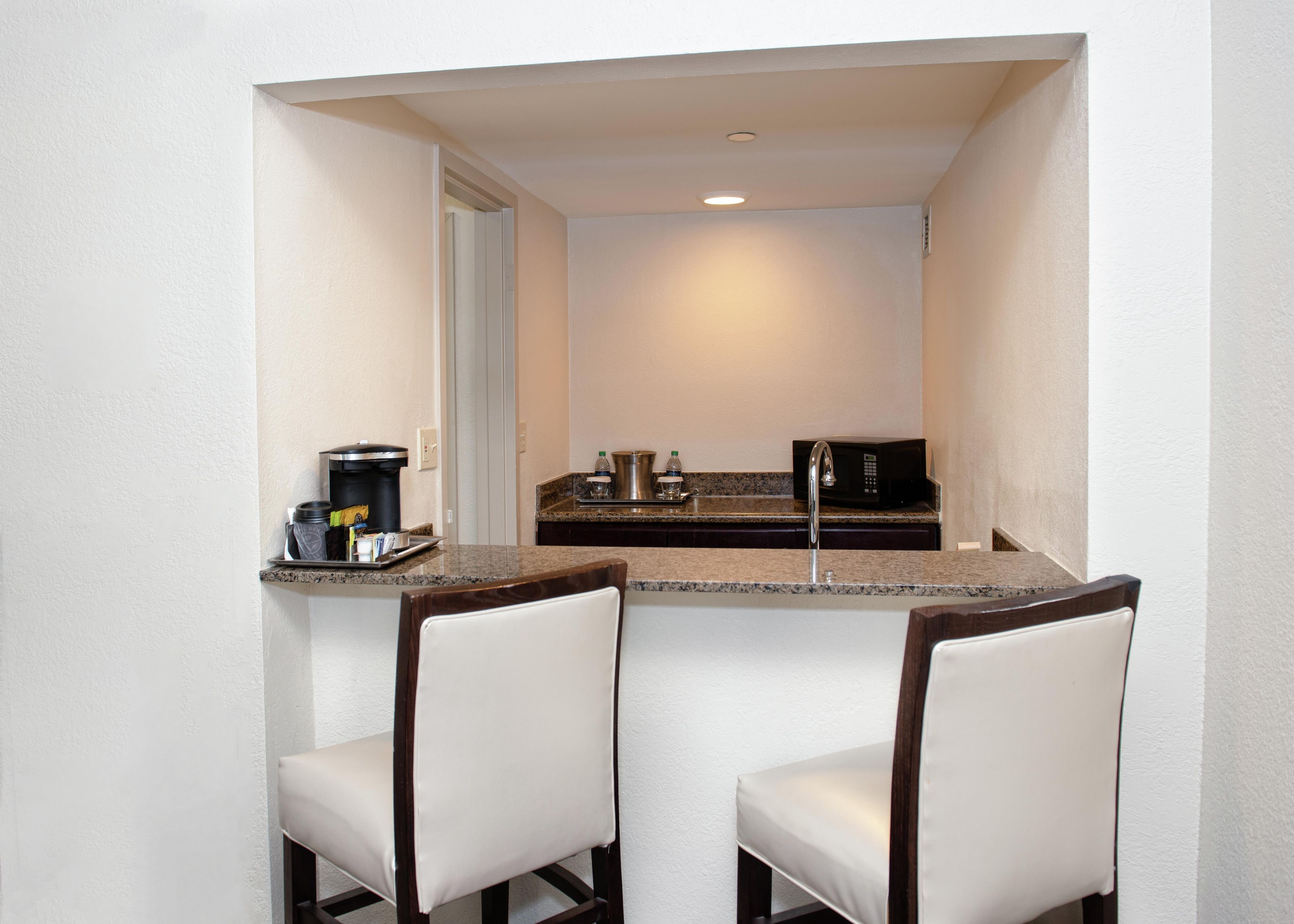 A small kitchenette with a microwave, coffee maker, and a sink. There are two white chairs in front of the counter.