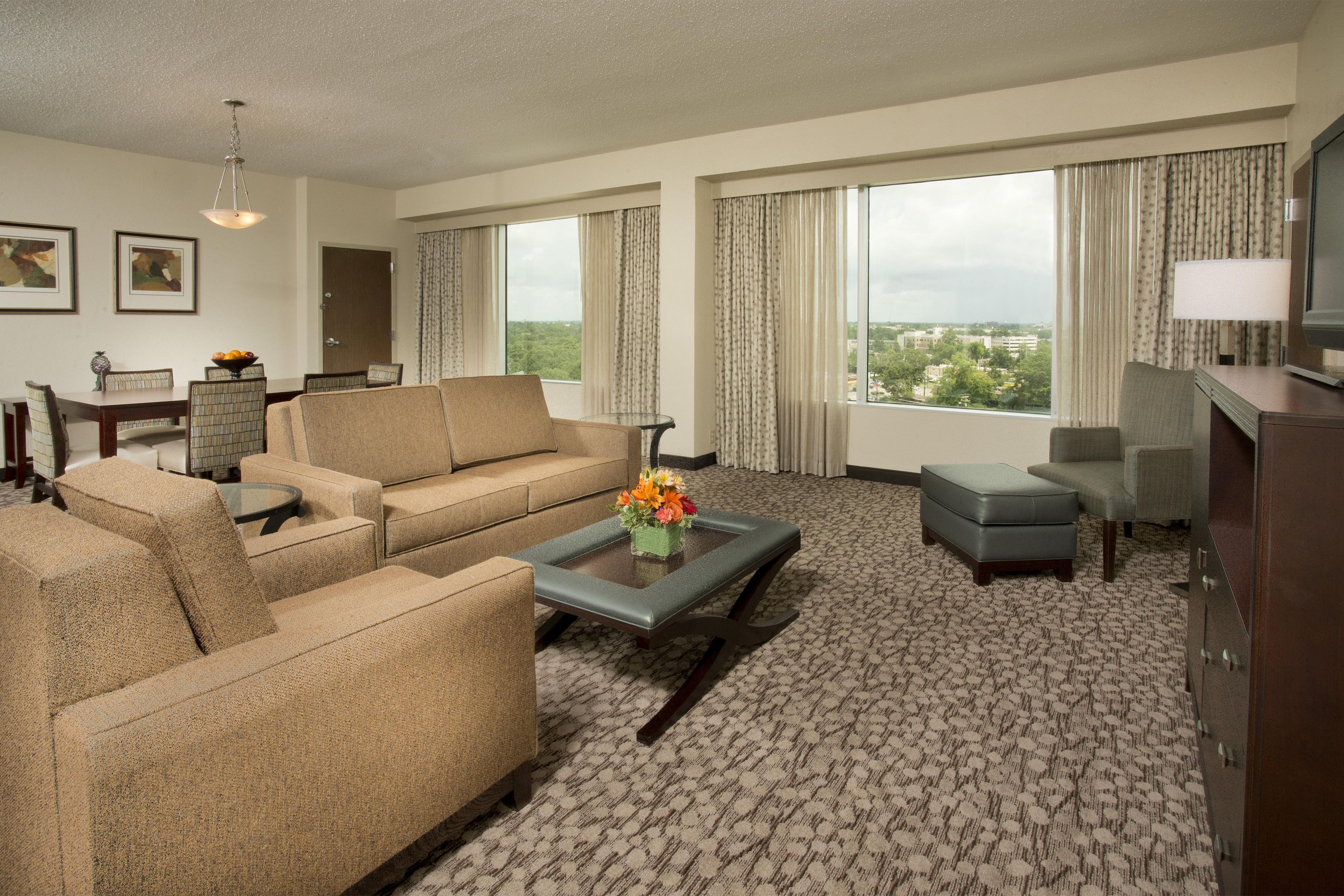 Un salón de hotel con un conjunto de sofás, una mesa de centro con flores, un televisor y vistas de la ciudad.