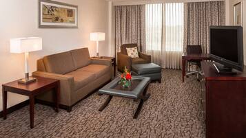 A hotel room with a sofa, armchair, coffee table, TV, and a desk.