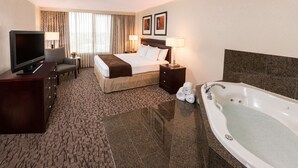 A hotel room with a bed, a TV, a desk, a chair, a desk lamp, a bedside table with a lamp, a bathtub, and a window with curtains.