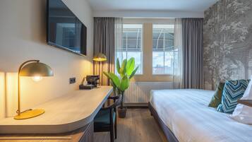 Ein modernes Hotelzimmer mit einer gebogenen Schreibtischplatte, einem Bett mit gemusterten Kissen, einem großen Fenster mit Vorhängen und einem an der Wand befestigten Flachbildfernseher.
