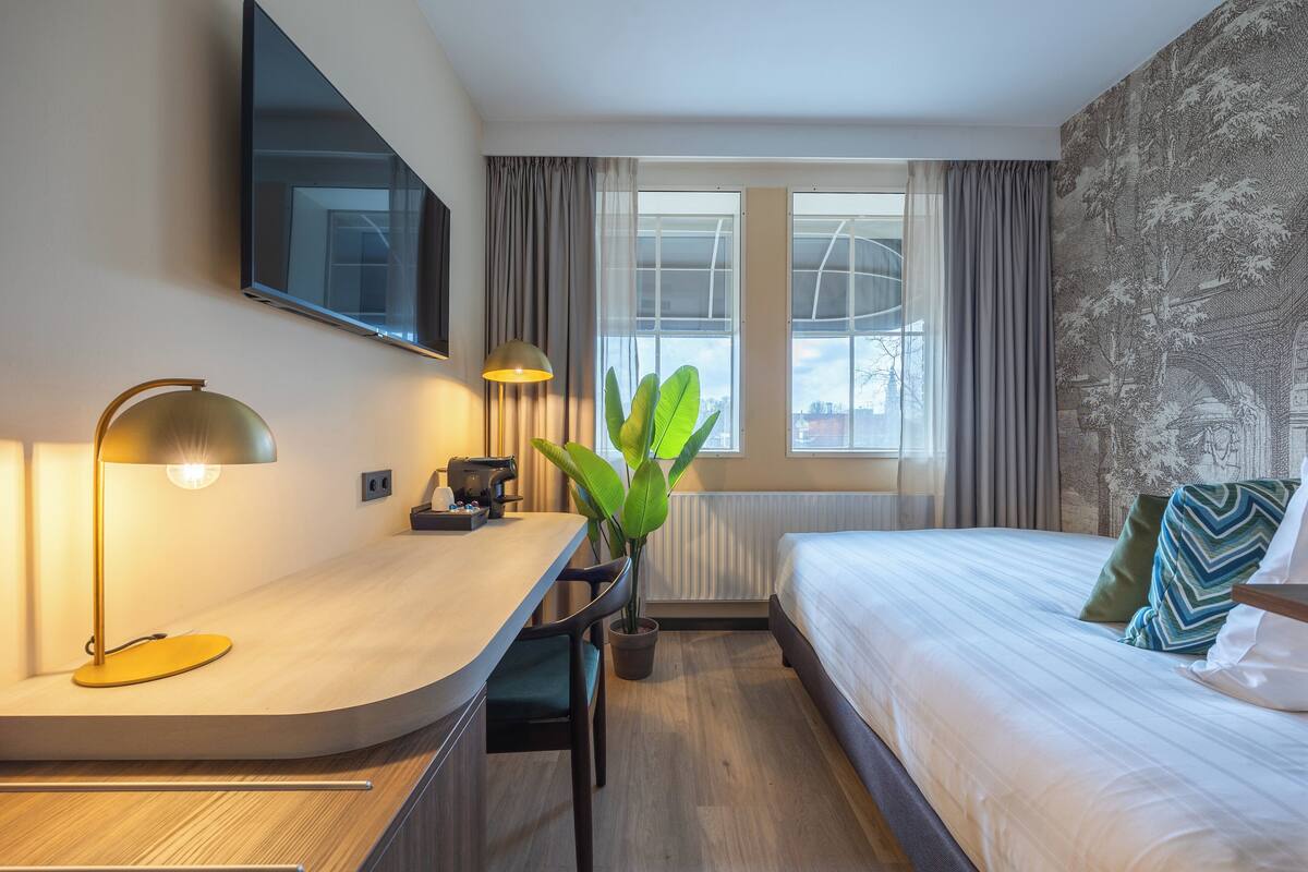 A modern hotel room with a curved desk, a bed with patterned pillows, a large window with curtains, and a mounted flat-screen TV.