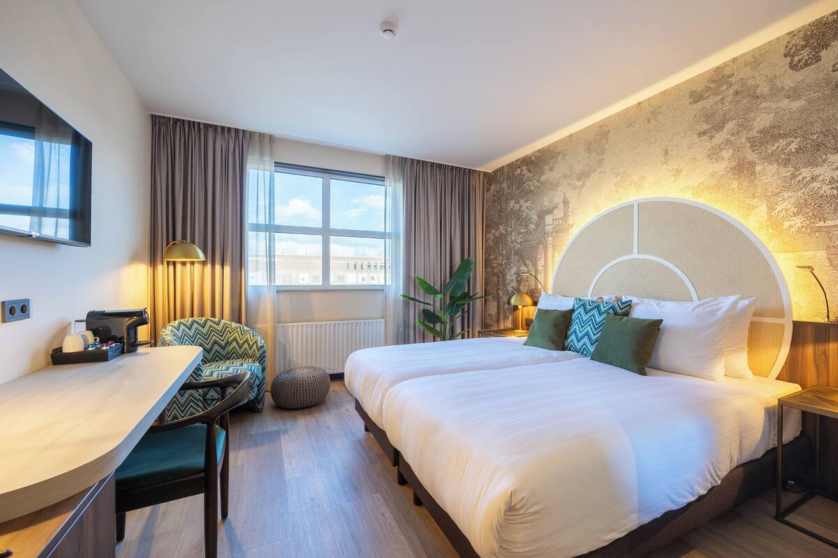 A modern hotel room with a large bed, a desk, and a chair. There is a window with curtains, a plant, and a wall-mounted lamp.