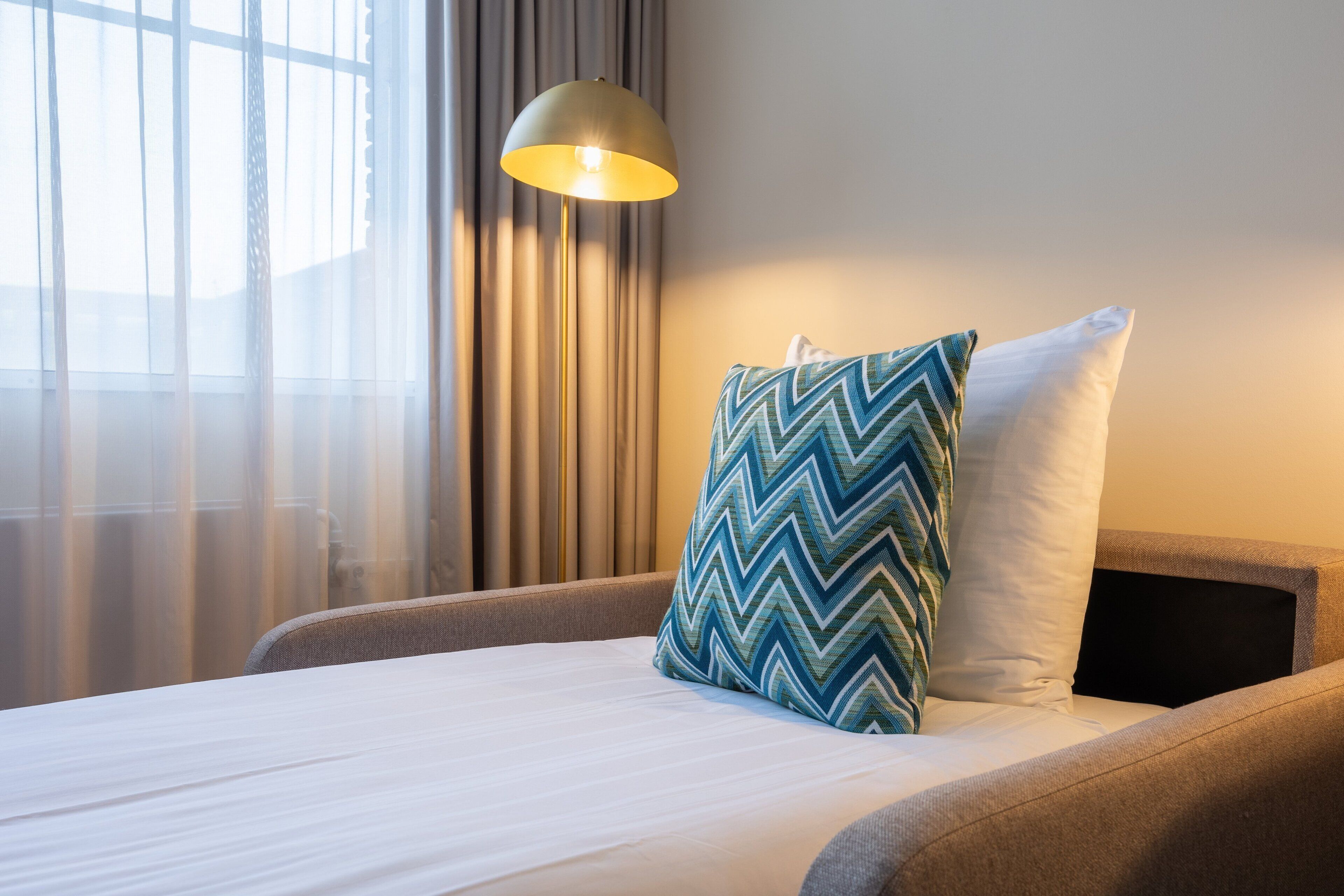 Una cama bien hecha con sábana blanca y almohada azul en zigzag, una lámpara de pie moderna y una ventana con cortinas de encaje.