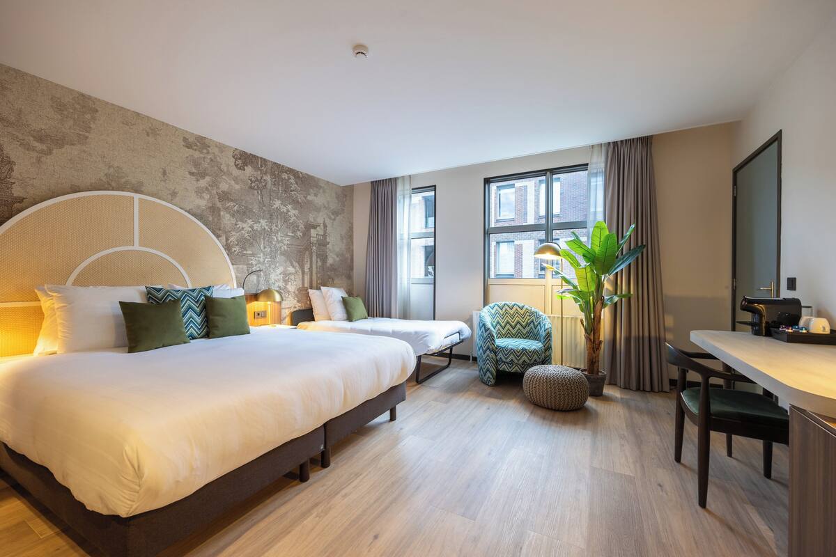 A hotel room with a large bed, two smaller beds, a desk, a chair, a potted plant, and a view of the city through large windows.