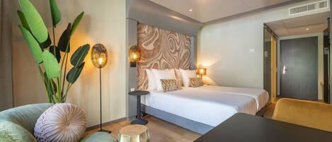 A modern hotel room with a bed, a sofa, a coffee table, and a large plant.