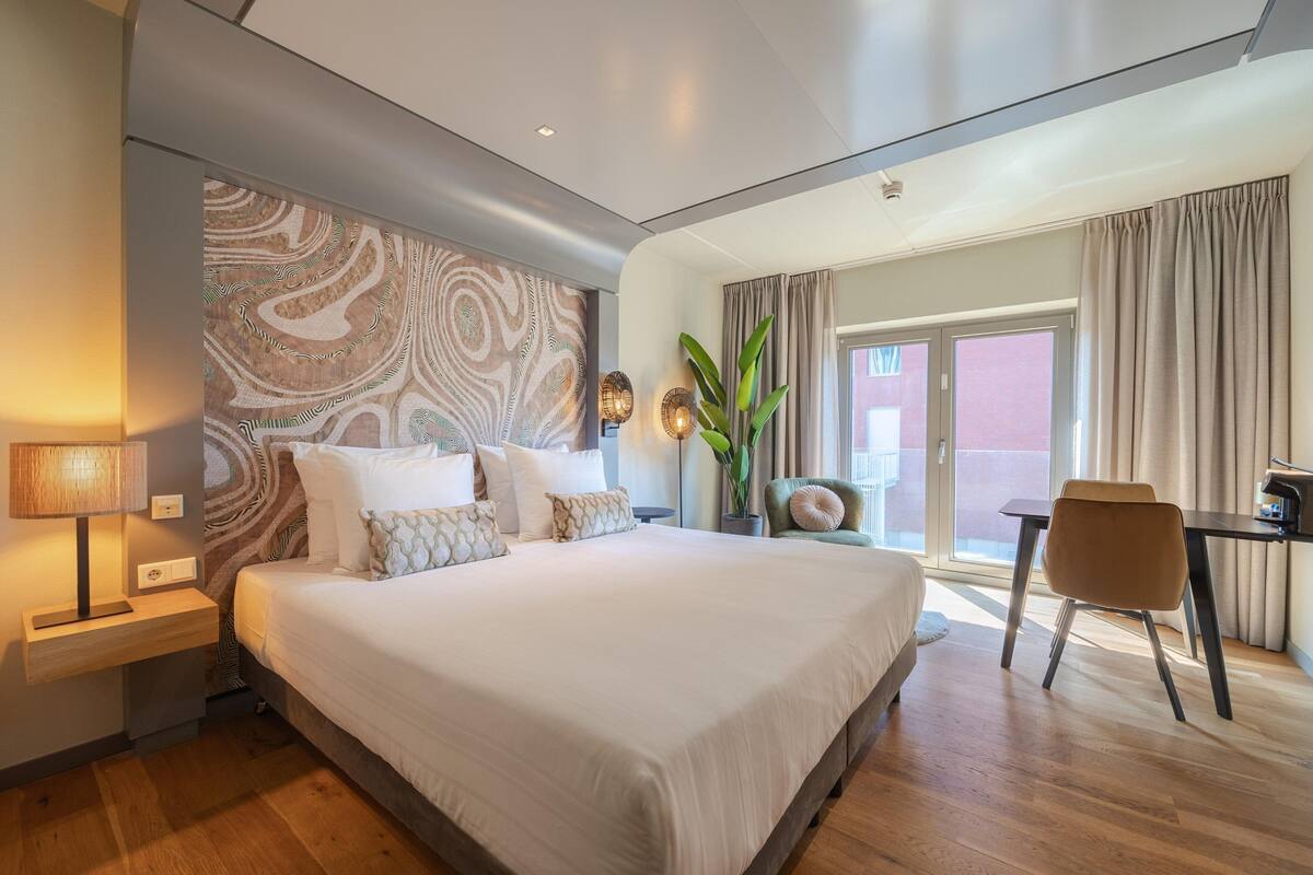 A modern hotel room with a large bed, a desk, and a view of the outdoors.