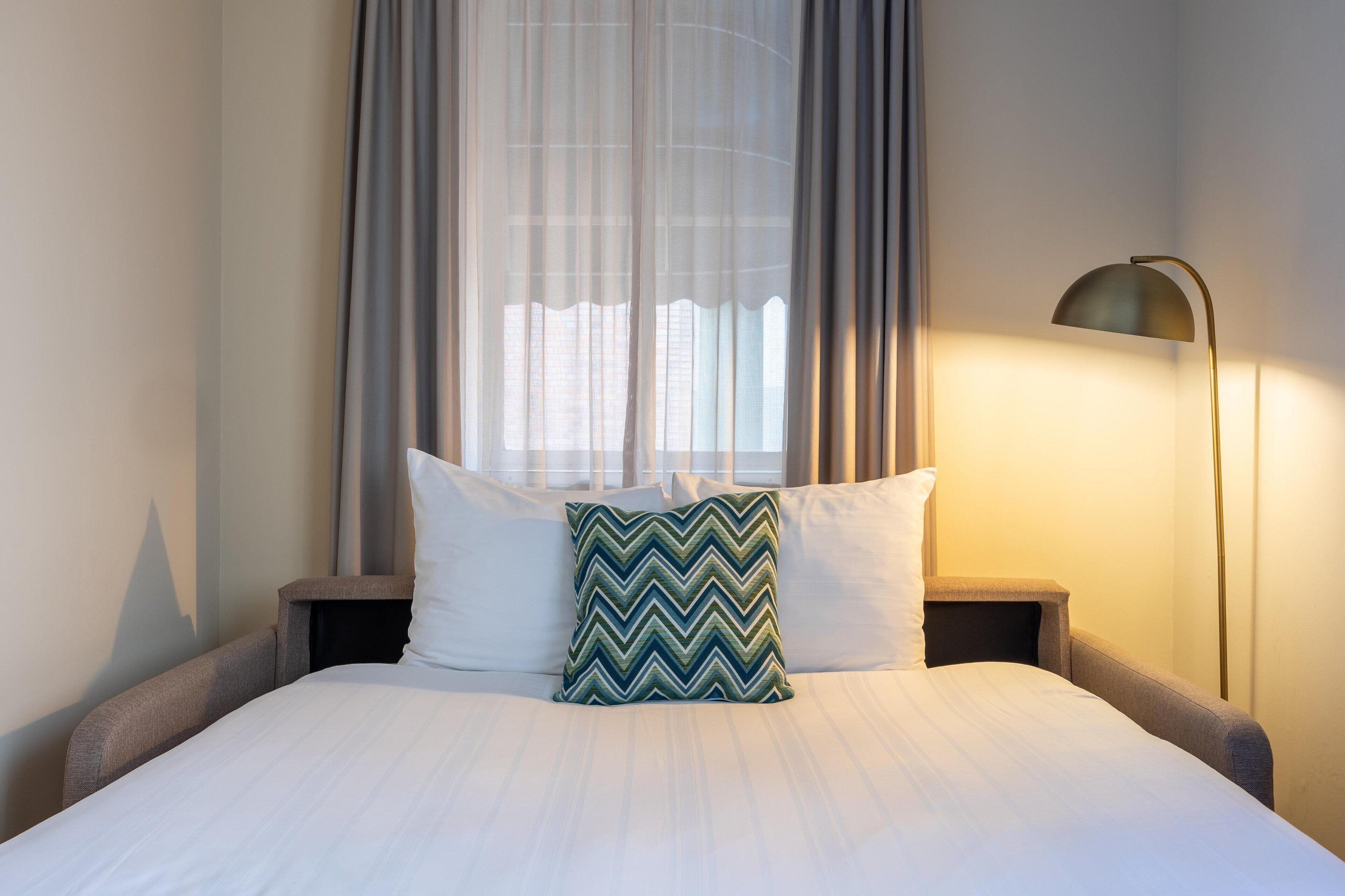Cama bem arrumada com roupa de cama branca e um travesseiro verde em zigue-zague, luminária de cabeceira e cortinas finas.