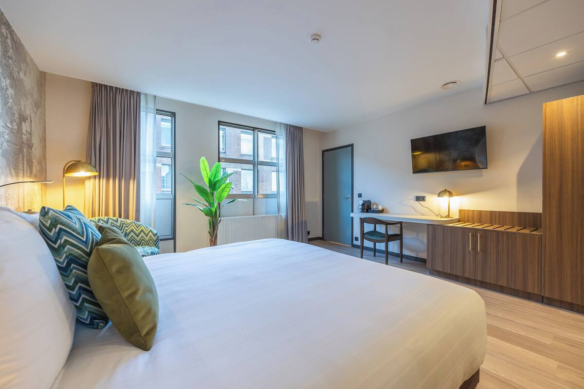 A hotel room with a large bed, a desk, a chair, a potted plant, and a TV on the wall.