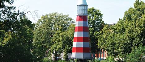 A conical lighthouse structure with a red and white striped pattern, situated on stilts over water, surrounded by lush greenery and trees.