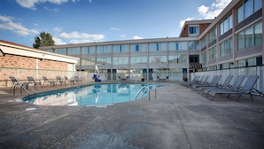 Une piscine entourée de chaises longues et de parasols, bordée d’un bâtiment doté de grandes fenêtres et d’une façade en briques.