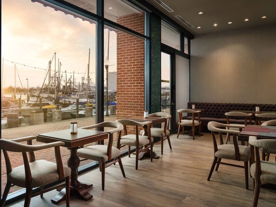 A restaurant with a view of a marina, featuring wooden tables and chairs, a brick wall, and large windows.