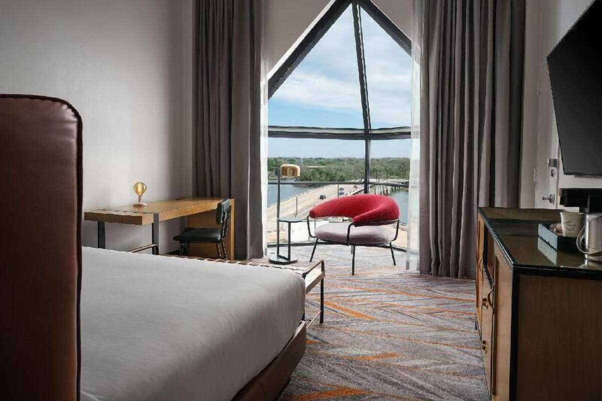 A hotel room with a bed, a desk, a chair, a TV, and a large window offering a view of a bridge and greenery.